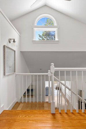 Un escalier avec des rampes blanches menant à un deuxième étage, une œuvre d’art encadrée accrochée au mur et une fenêtre donnant sur des arbres.