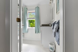 Une salle de bain avec un sol carrelé de carreaux blancs, une fenêtre avec des rideaux, un lavabo avec un robinet et des serviettes suspendues sur un porte-serviettes.