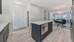 A modern kitchen with a central island, a dining area with a table and chairs, and a view of the city through large windows. - A Family Relaxing Vacation House, 3 bedrooms2.5 bathrooms for up to 8 people (Calgary)