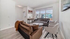 A modern living room with a brown sofa, grey couch, and white armchairs. There is a coffee table and a TV mounted on the wall. - A Family Relaxing Vacation House, 3 bedrooms2.5 bathrooms for up to 8 people (Calgary)