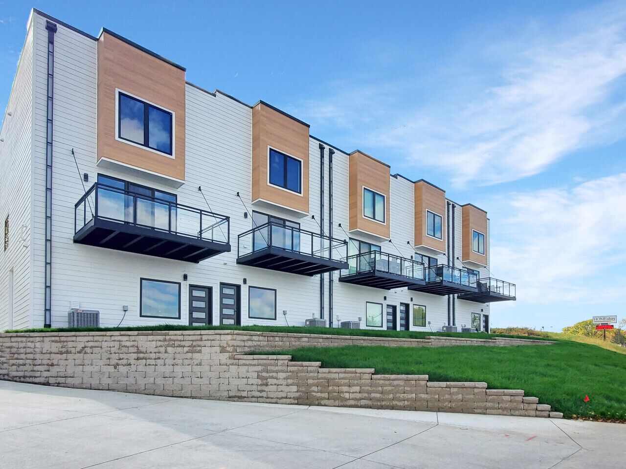 A modern multi-story residential building with balconies, a brick foundation, and a grassy area in front.