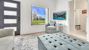Ein modernes Wohnzimmer mit grauem Sofa, pouf mit Grau-Steppung, grauem Sessel, großem Fenster mit Blick auf eine grüne Landschaft und einem gerahmten Gemälde an der Wand.