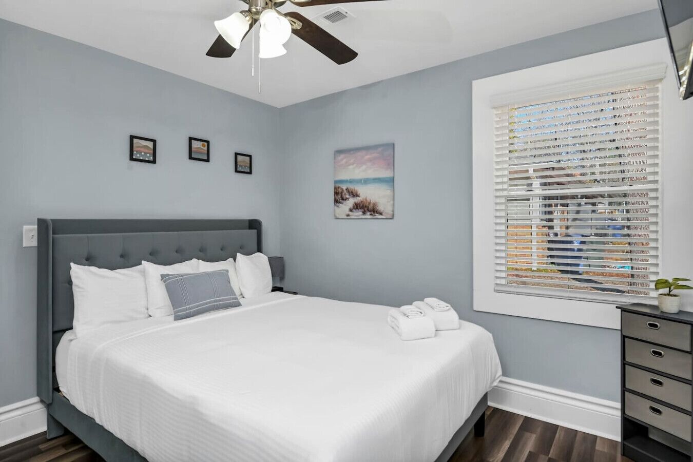 Une chambre à coucher avec un lit, un ventilateur de plafond, une fenêtre avec des stores et une petite plante posée sur une commode.