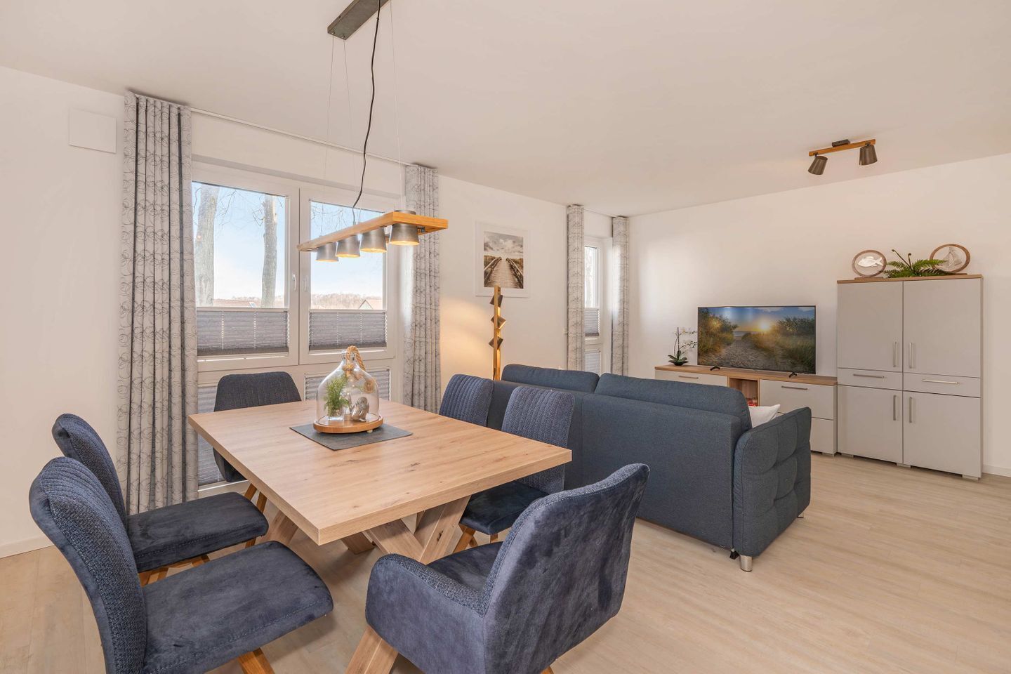 A modern living room with a wooden dining table, blue chairs, a grey sofa, a TV, and a cabinet.