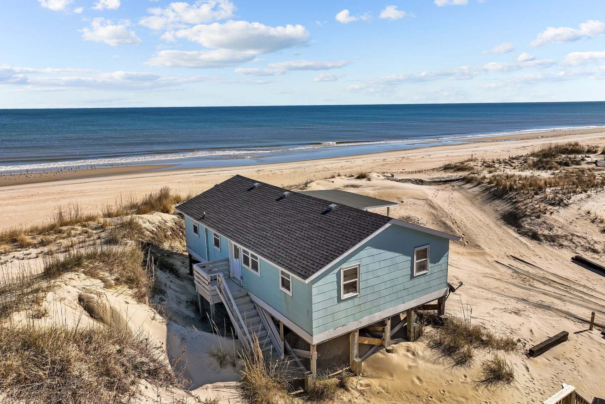 A beach house with a staircase leading to a deck, situated on sandy terrain with dunes and a view of the ocean.