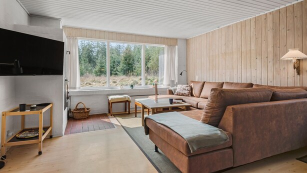 A modern living room with a large window, wooden walls, a brown leather sofa, a small table, and a floor lamp. - Cozy home in Nørre Nebel with sauna (Nørre Nebel)