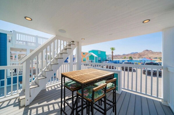 Une terrasse en bois avec une table et des chaises, une balustrade blanche, et une vue sur des bâtiments aux couleurs vives et une montagne en arrière-plan.