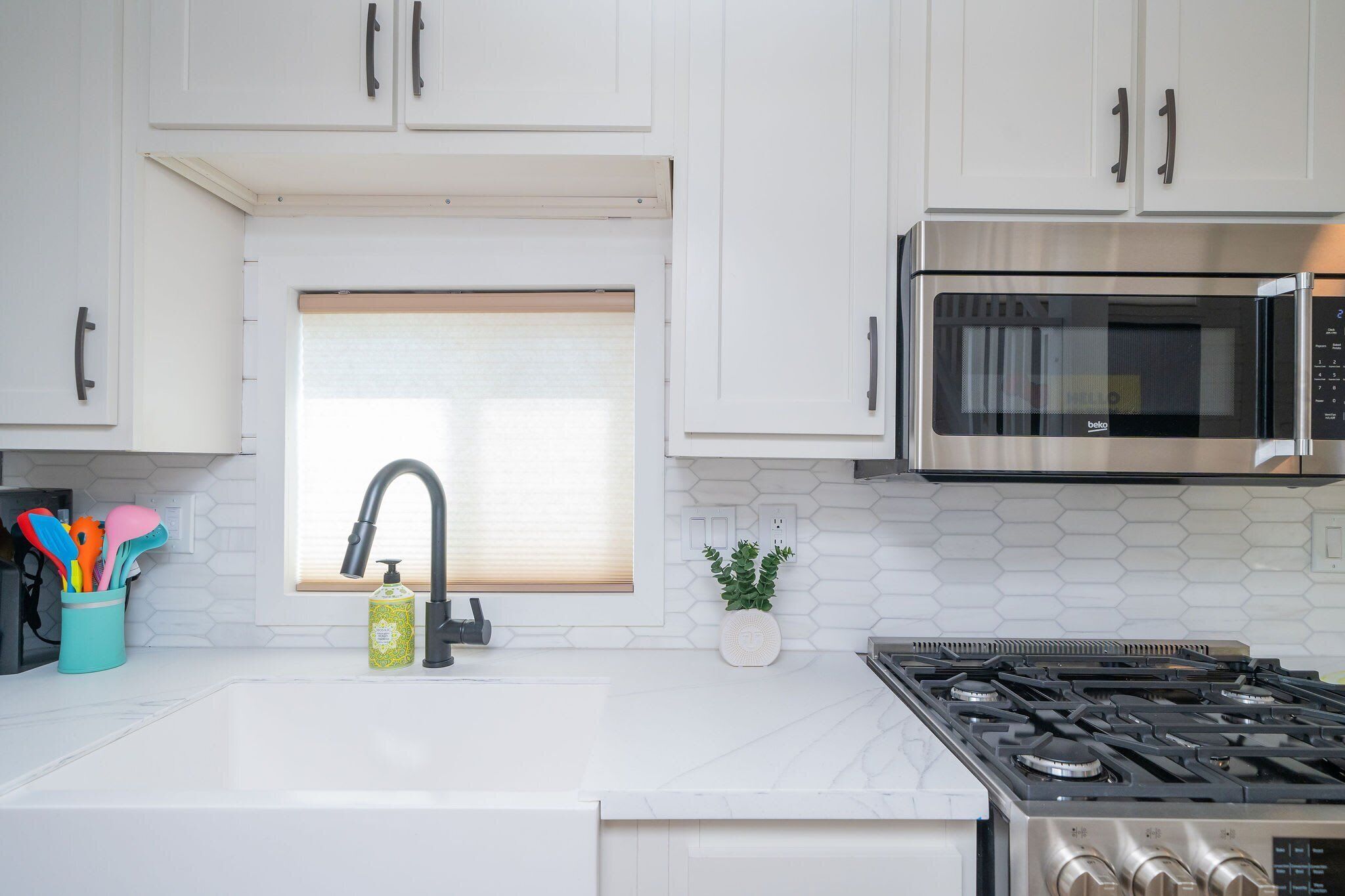 A modern kitchen with white cabinets, a stainless steel microwave, a gas stove, a window with blinds, and a small potted plant.