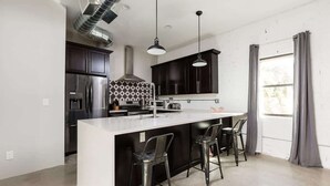 A modern kitchen with a central island, bar stools, dark cabinetry, stainless steel appliances, and a large window with curtains. - Nova-400k Backyard! Old Town and Ent District! (Scottsdale)