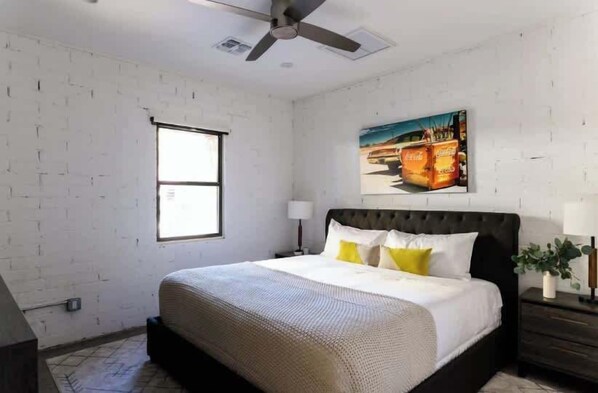 Un dormitorio con cama, ventilador de techo, ventana y un cuadro en la pared.