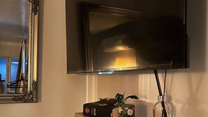 A modern living room with a mounted flat-screen TV, a decorative mirror, and a wooden shelf with various items including a remote control, a decorative bowl, and a small vase. - Beach Serenity 1

Share

Save
 (Brighton-Le-Sands)