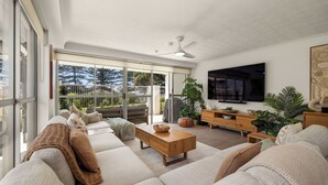 A spacious living room with a large sofa, a wooden coffee table, and a TV mounted on the wall. There is a sliding glass door leading to a balcony with a view of trees and a fence.