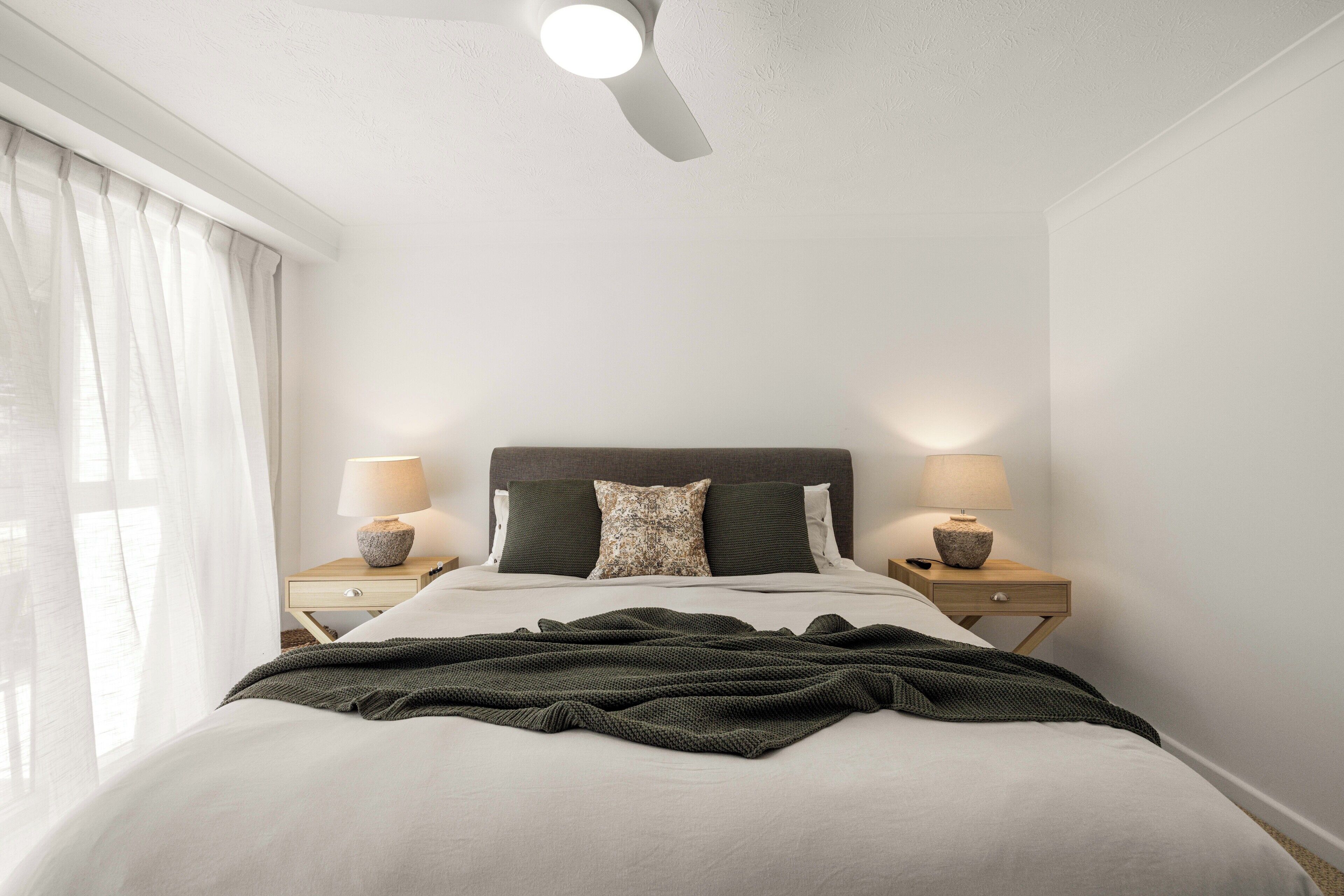 A neatly made bed with a dark green blanket, two bedside lamps, and a ceiling fan in a simple, clean room with white walls and a window with sheer curtains.