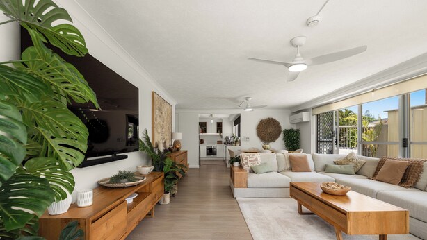 A modern living room with a sofa, coffee table, and TV. There are plants and a kitchen in the background.
