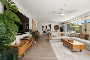 A modern living room with a sofa, coffee table, and TV. There are plants and a kitchen in the background. - Cashelmara on the Beach 3 Bedroom Escape (Burleigh Heads)