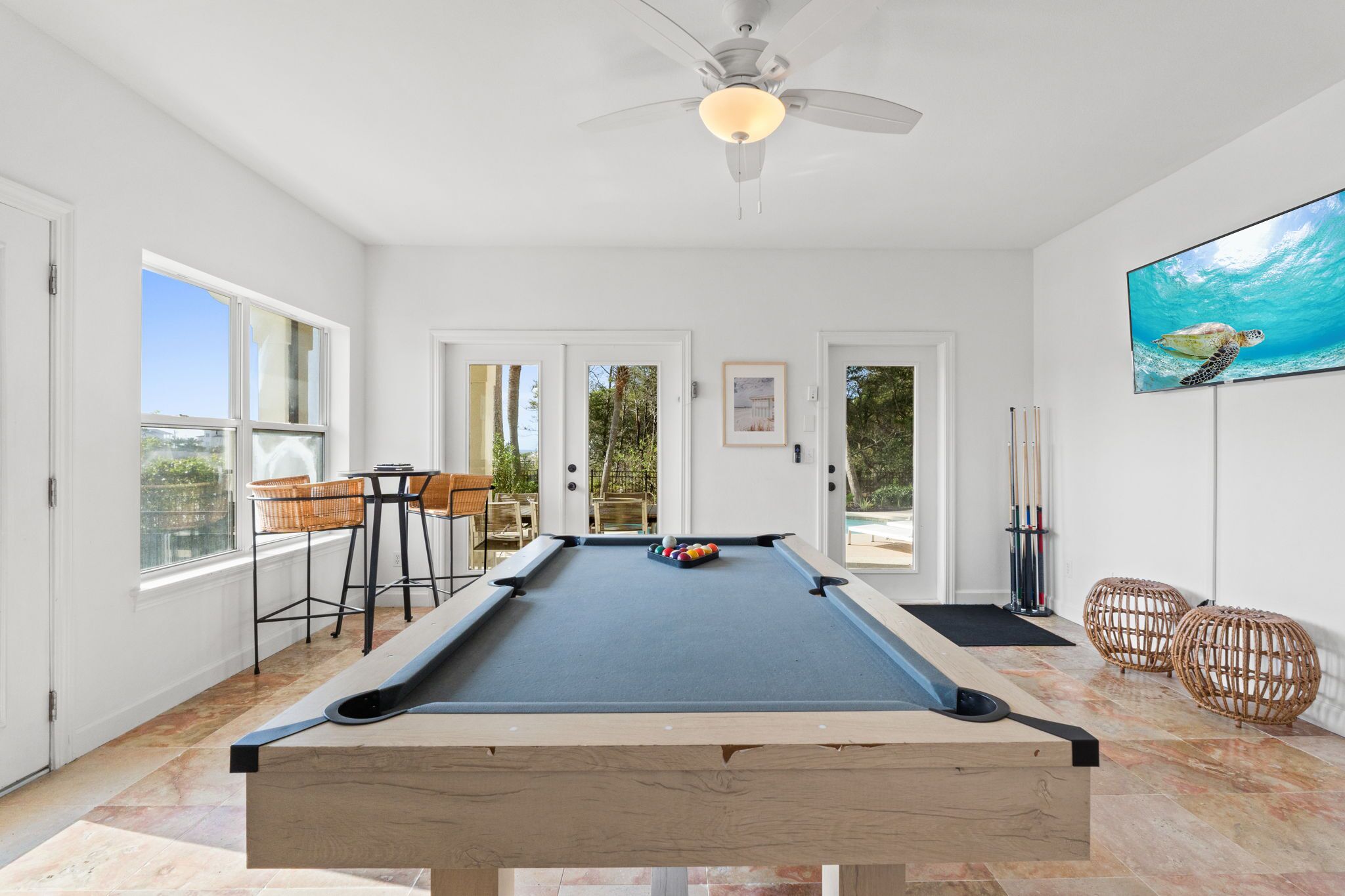 A pool table with a ceiling fan, a television, and a pool cue rack in a bright room with large windows.