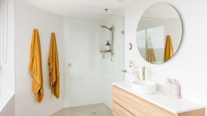 A modern bathroom with a glass shower enclosure, a round mirror, and a white vanity with a sink and storage. - Ocean View Coastal Haven in Burleigh (Burleigh Heads)