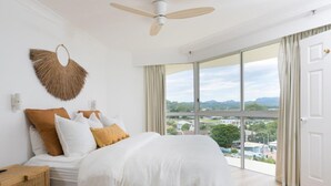 A bedroom with a large bed, a ceiling fan, a view of the city through large windows, and a door to the right. - Ocean View Coastal Haven in Burleigh (Burleigh Heads)