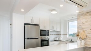 A modern kitchen with white cabinetry, stainless steel appliances, a large island, and a view through the window. - Ocean View Coastal Haven in Burleigh (Burleigh Heads)