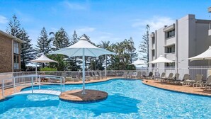 A swimming pool with lounge chairs and umbrellas, surrounded by a white fence and trees. - Ocean View Coastal Haven in Burleigh (Burleigh Heads)