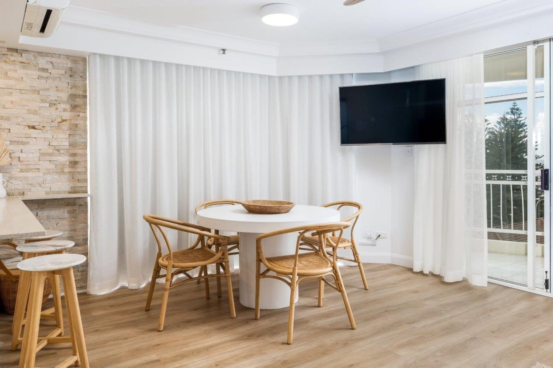 A modern living room with a round table, four wicker chairs, a flat-screen TV mounted on the wall, and a large window with white curtains.