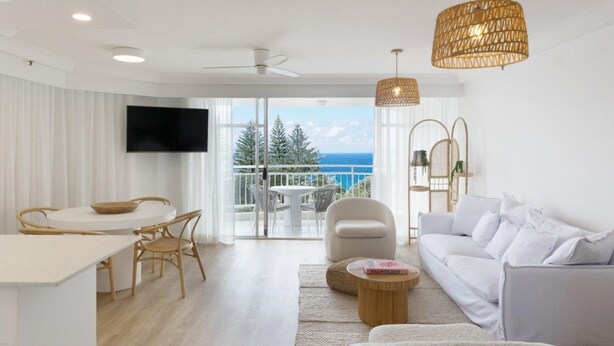 A modern living room with a white sofa, a dining table with chairs, a TV mounted on the wall, and a balcony with a view of the ocean. - Ocean View Coastal Haven in Burleigh (Burleigh Heads)