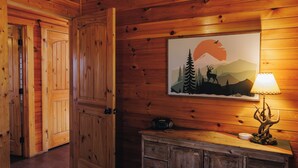 Interior de una cabaña de madera con una puerta grande, un cuadro de un paisaje montañoso, una lámpara y un armario.