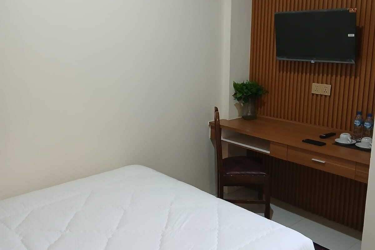 Hotellihuone, jossa on yksi sänky, puinen työpöytä tuolilla, seinälle asennettu televisio ja pieni ruukkukasvi.