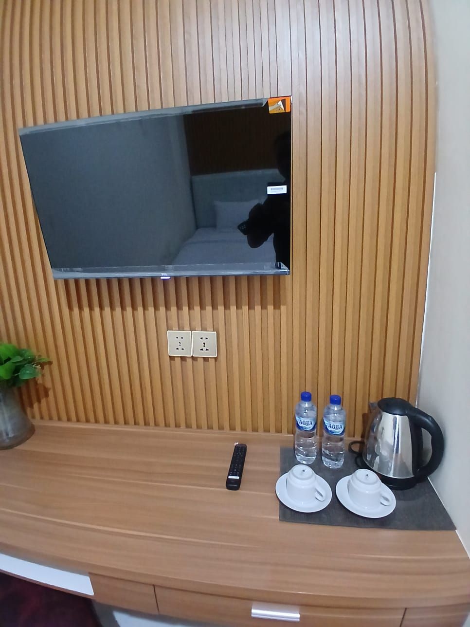 A hotel room with a wooden panel wall, a flat-screen TV mounted above a wooden desk, a potted plant, a remote control, two water bottles, a kettle, and two white teacups.