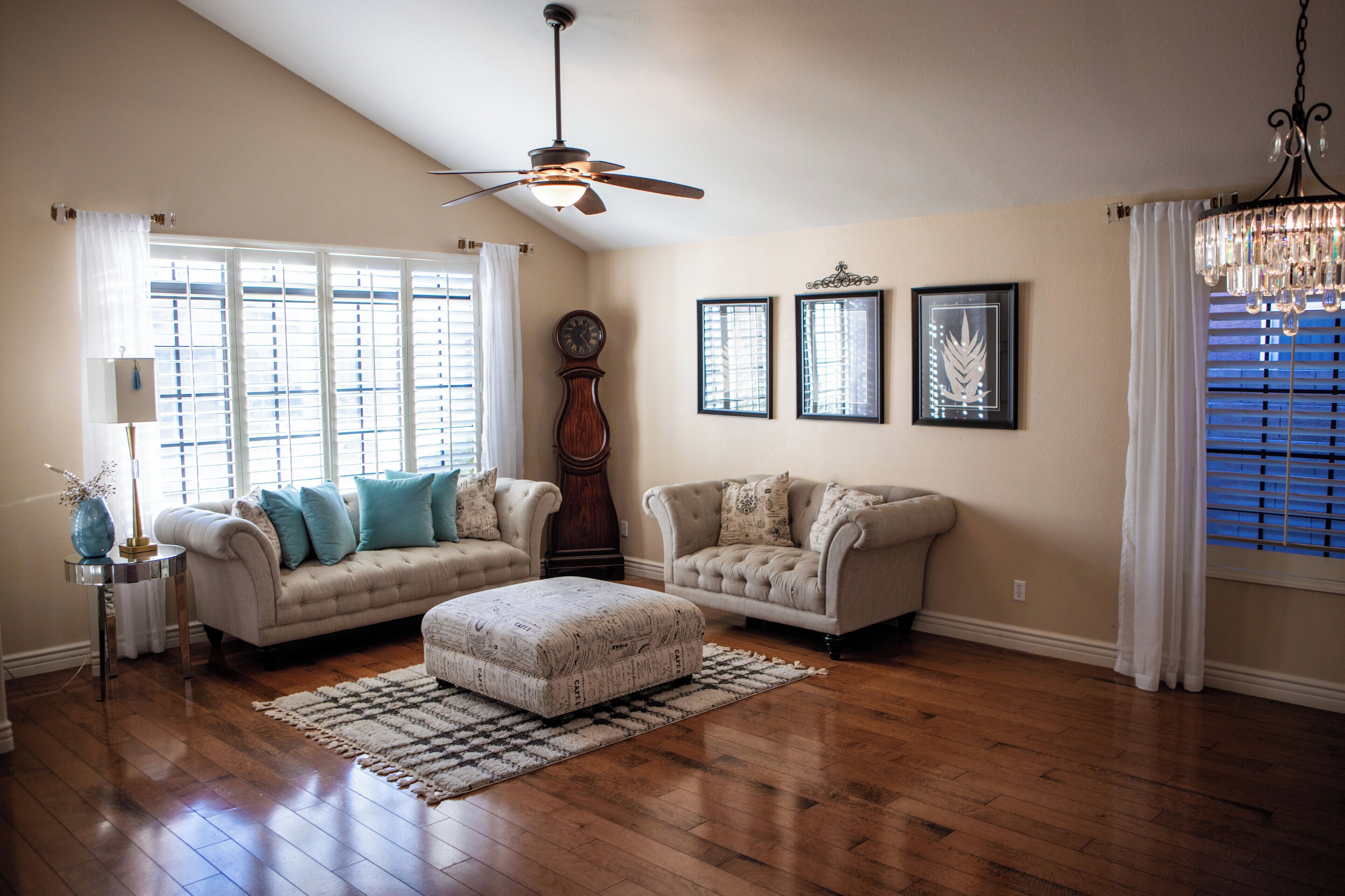 A spacious living room with hardwood flooring, two sofas, a large ottoman, a ceiling fan, and a chandelier.