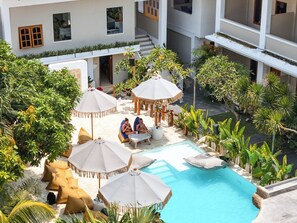 A pool area with lounge chairs and umbrellas, surrounded by greenery and a building with multiple floors. - Mad Monkey Uluwatu (Pecatu)