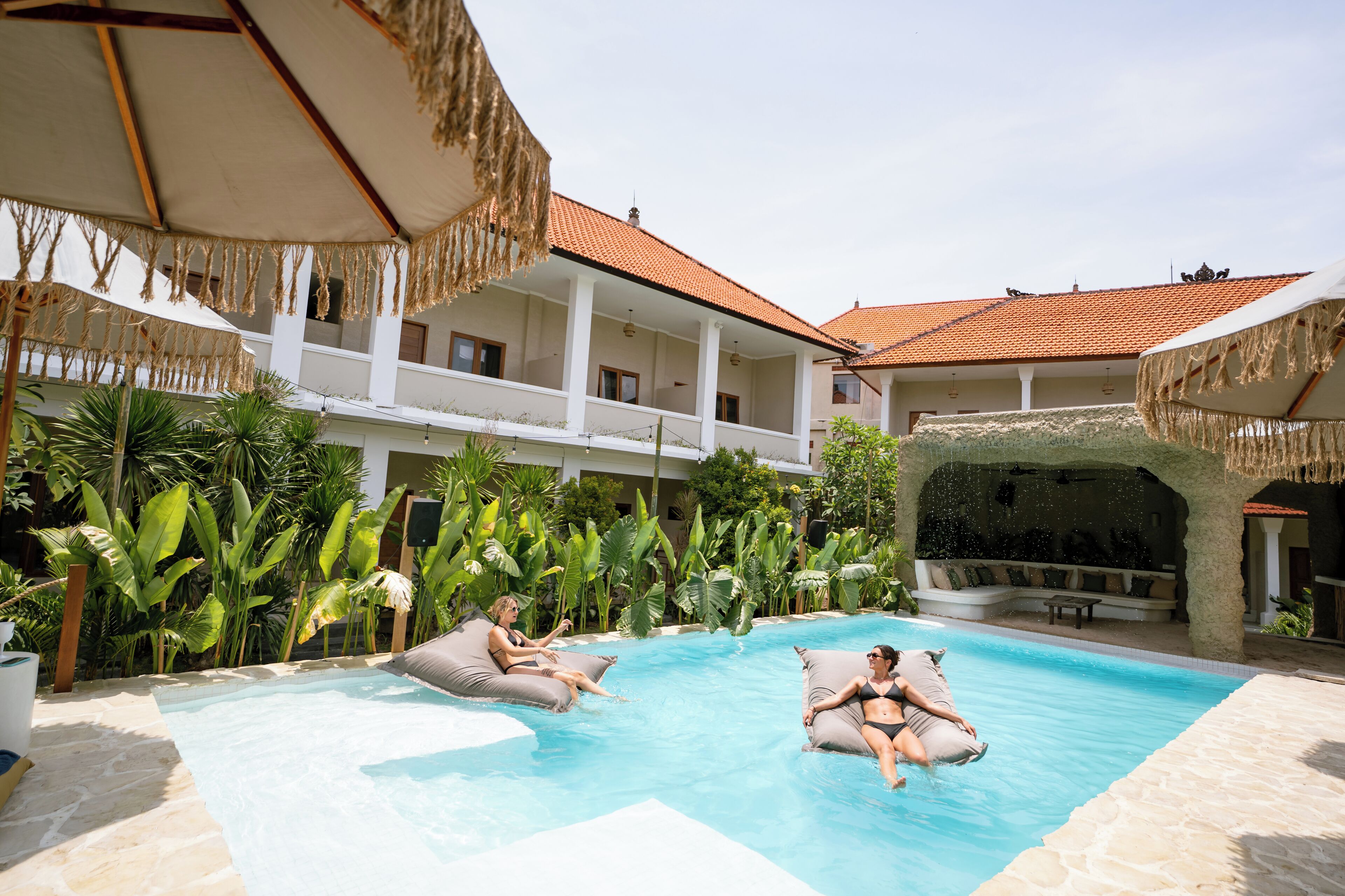 Uma área de piscina com espreguiçadeiras e guarda-sóis, cercada por vegetação exuberante e um edifício com telhado de azulejos.