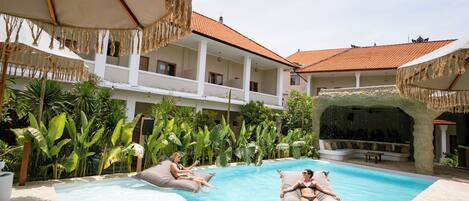 Uma área de piscina com espreguiçadeiras e guarda-sóis, cercada por vegetação exuberante e um edifício com telhado de azulejos.