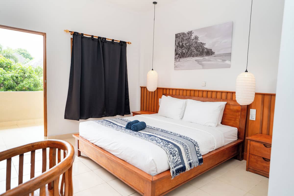 A bedroom with a wooden bed, white bedding, a black curtain, a wooden nightstand, and a white wall with a picture.