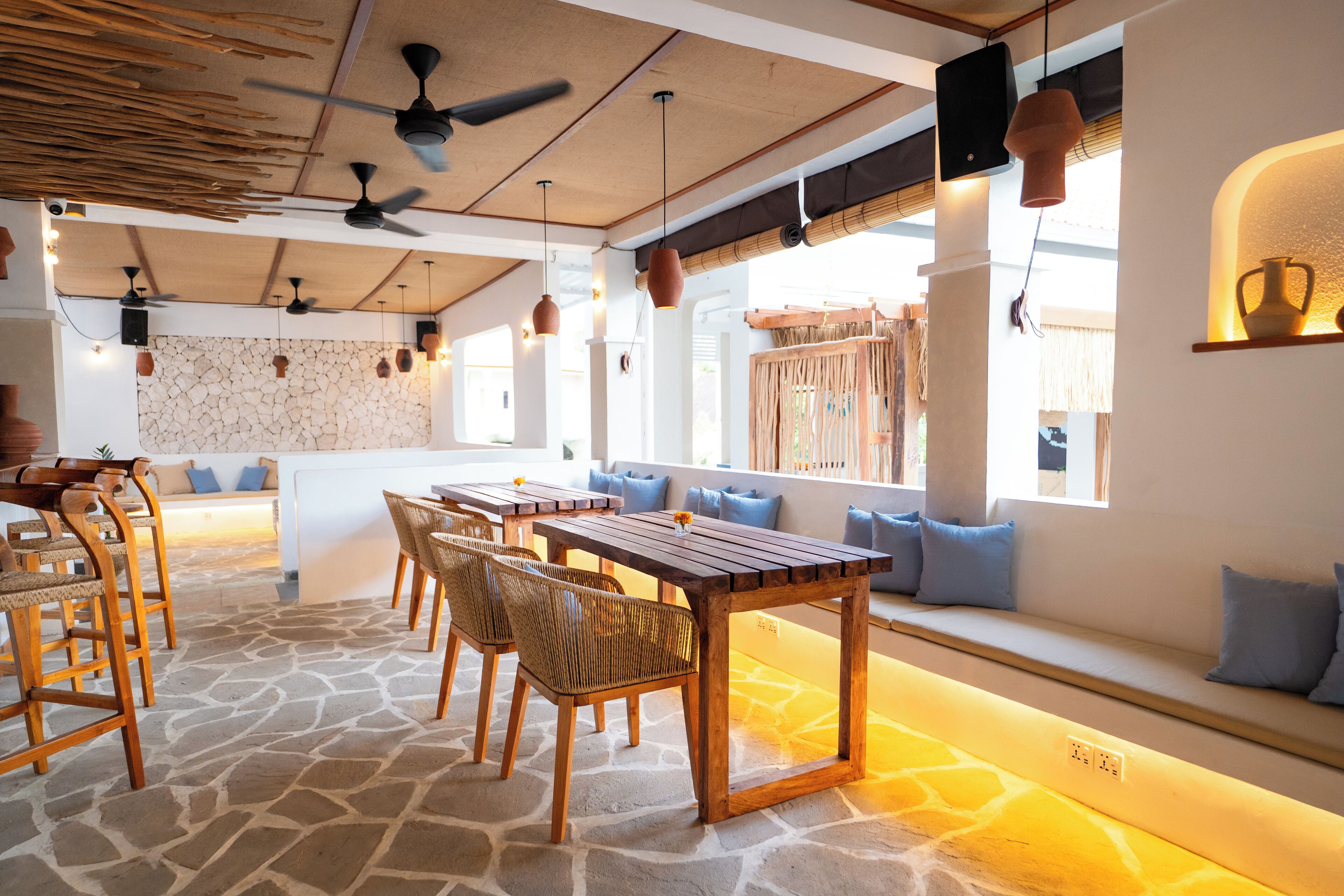 A restaurant with a stone floor, wooden tables, and chairs. There are ceiling fans and pendant lights.