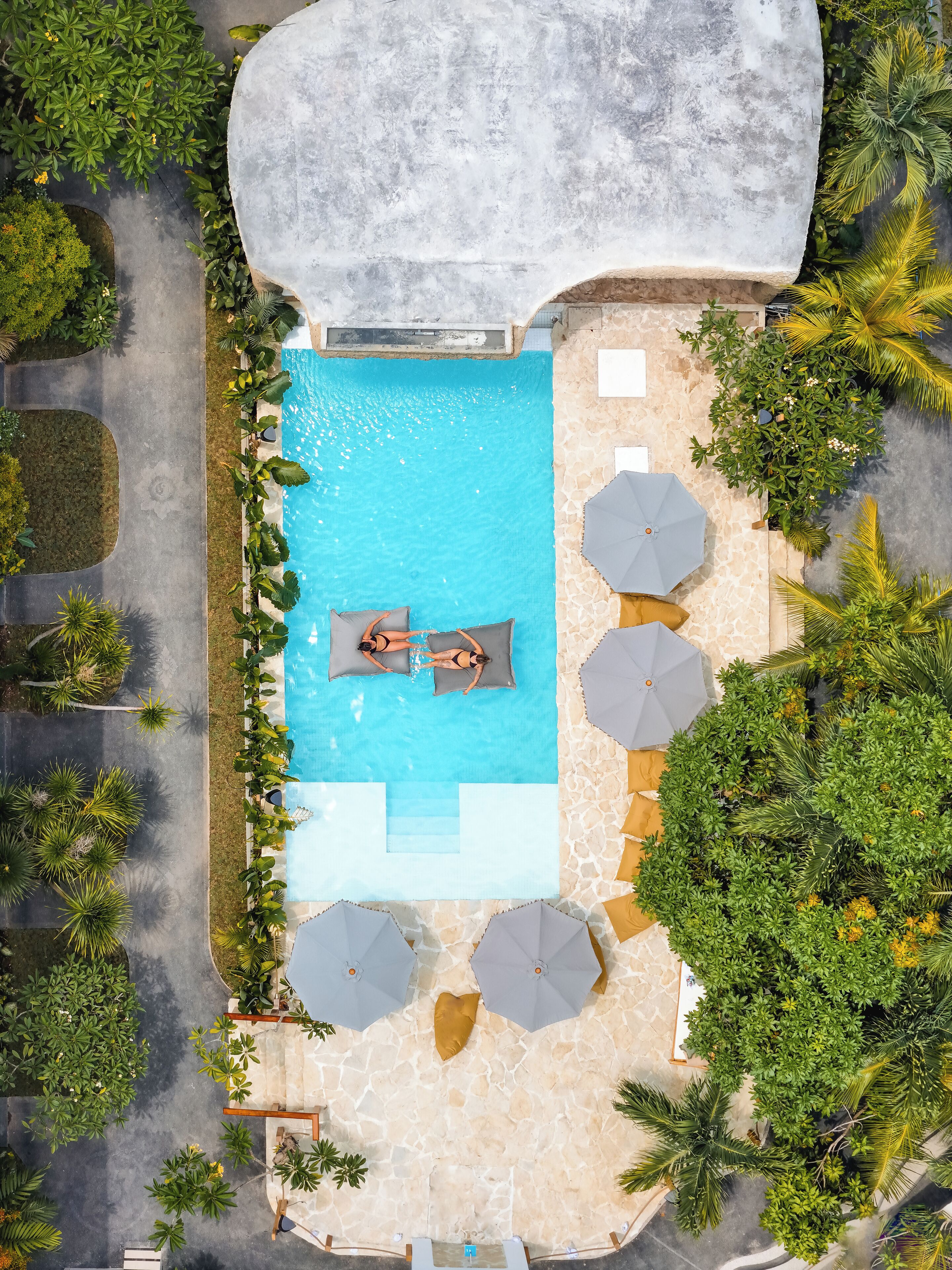 An aerial view of a pool area with two lounge chairs, umbrellas, and surrounding greenery.