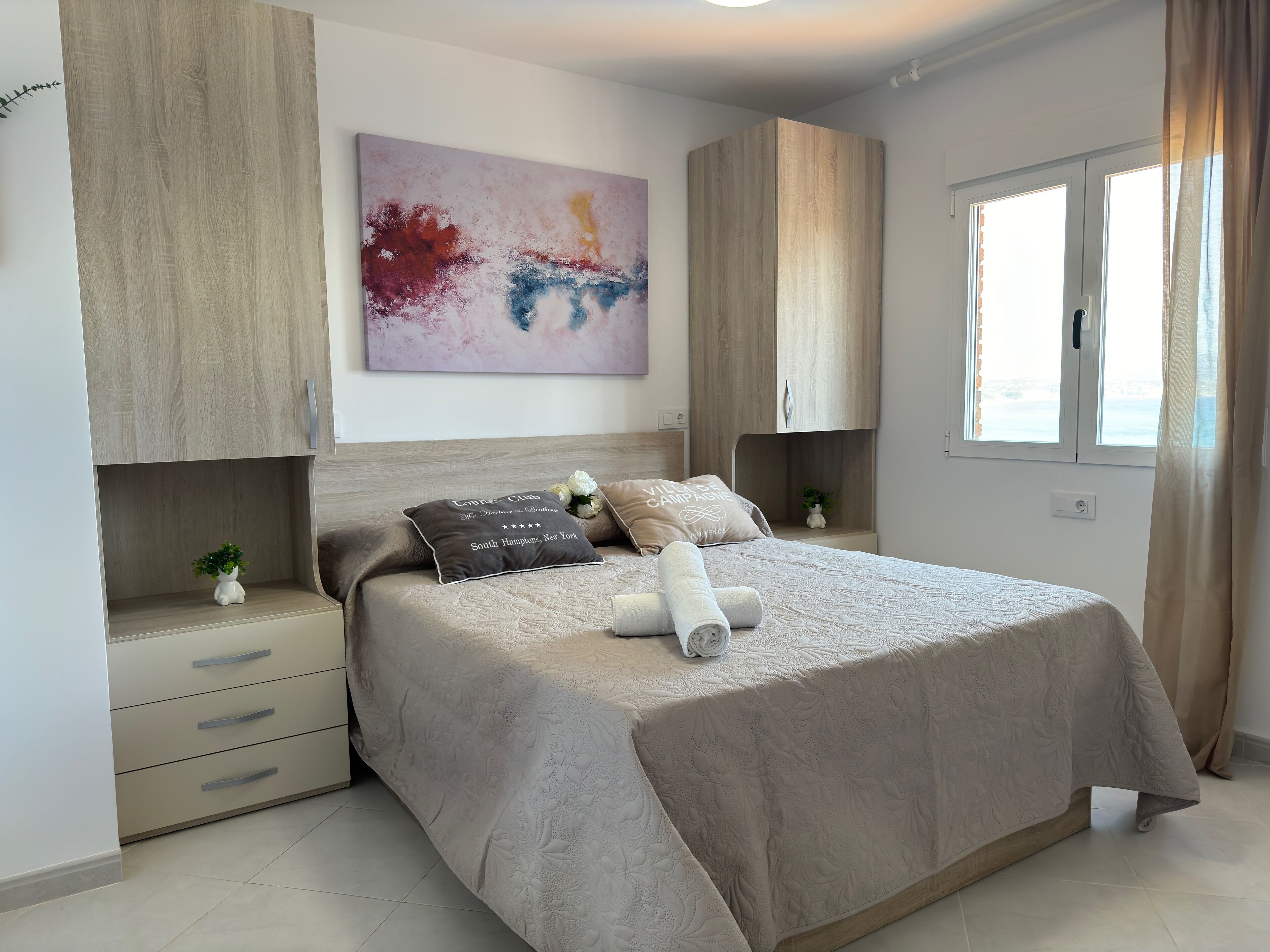 Un dormitorio moderno con una cama grande, un armario de madera, una mesita de noche con una planta y un cuadro en la pared.