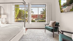 Ein Schlafzimmer mit einem großen Bett, einer Sitzecke mit einem Sessel, einem an der Wand montierten Fernseher und einem Fenster mit Blick auf einen Pool und Grünflächen.