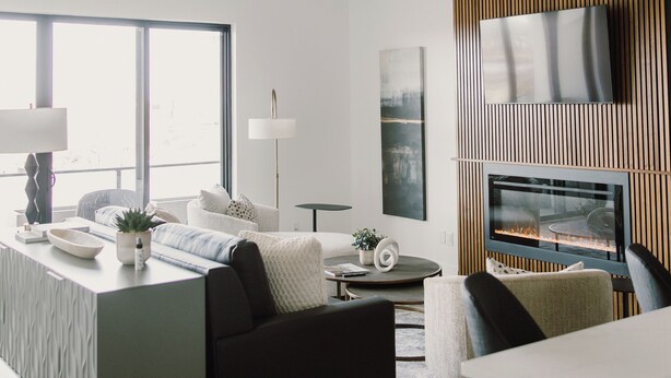 A modern living room with a large window, a flat-screen TV mounted on a wooden panel, a ceiling fan, and a dining area with a table and chairs. - Modern Townhome with Skyline Views, Heated Balcony & Curated Art in Downtown DSM (Des Moines)