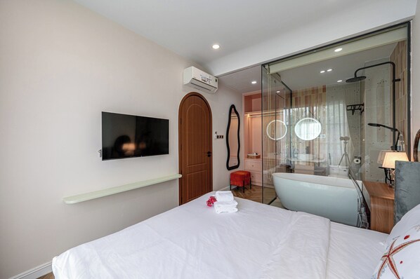 Habitación de hotel con cama, televisión, espejo, zona de ducha y un pequeño espacio de descanso.