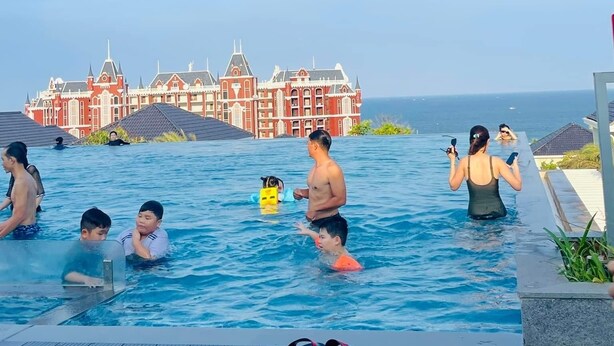 A swimming pool with people, a large building in the background, and a clear sky. - Venus Villa - Novaworld Phan Thiet (Tien Thanh)
