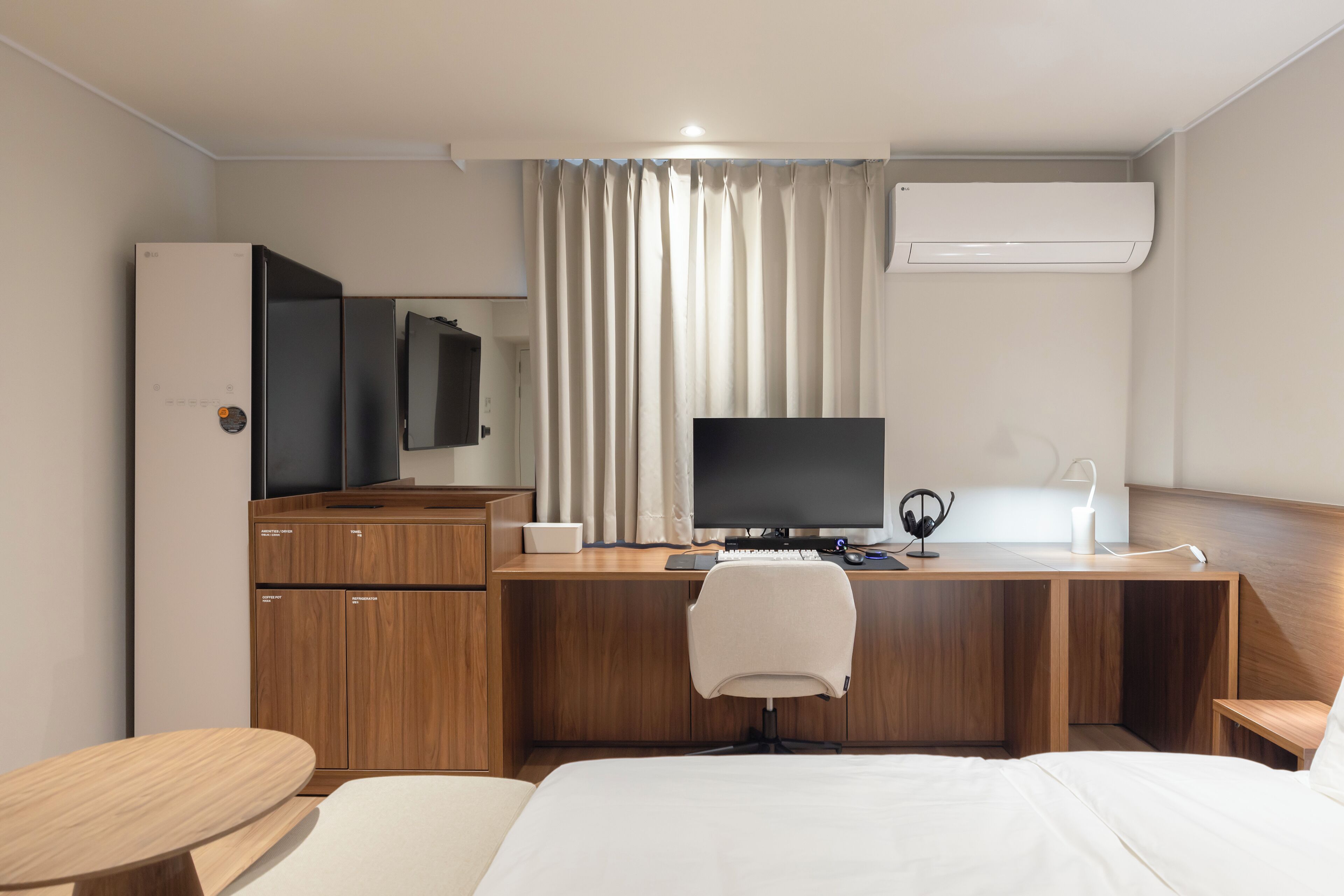 A hotel room with a wooden desk, a bed, a TV, a refrigerator, and an air conditioning unit.