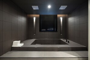 A modern bathroom with a rectangular bathtub, two wall-mounted lights, and a stack of white towels on the floor. - Anook Hotel Yeongdengpo (Seoul)