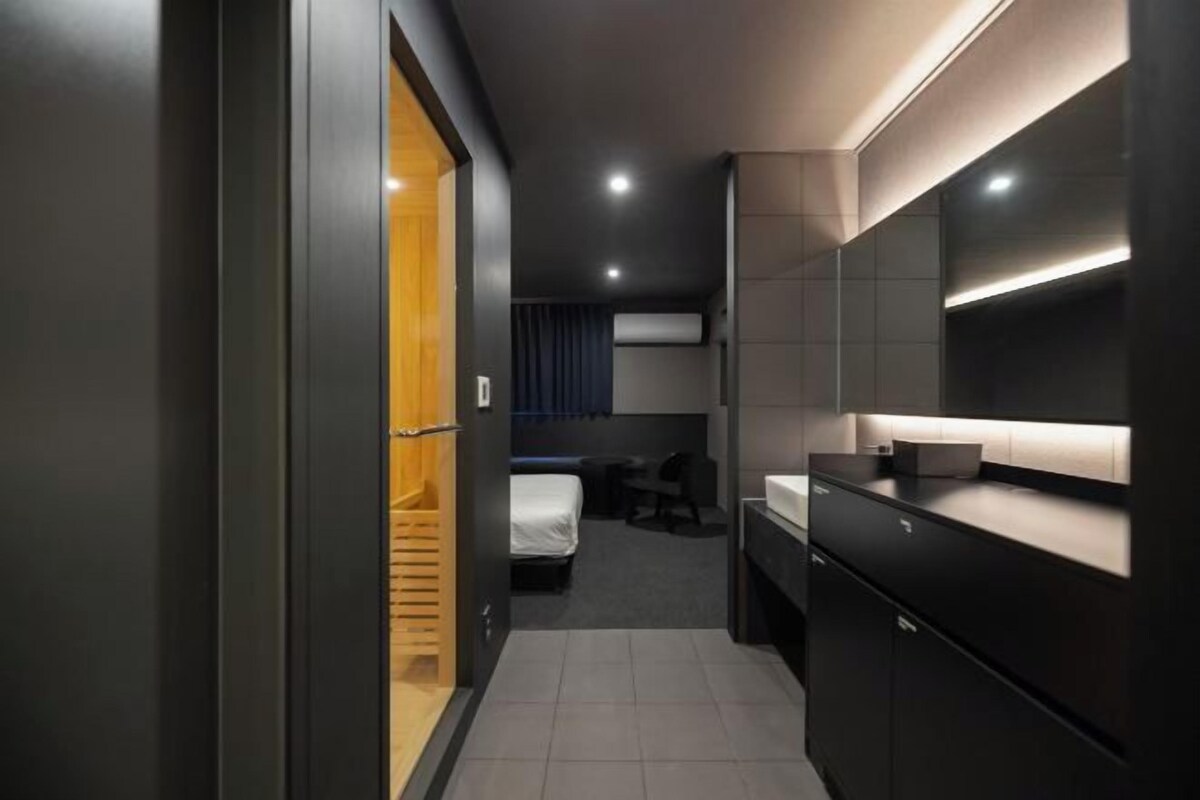 Una habitación de hotel moderna con una decoración en tonos oscuros, un armario de madera, una cama, un escritorio y un espejo grande.
