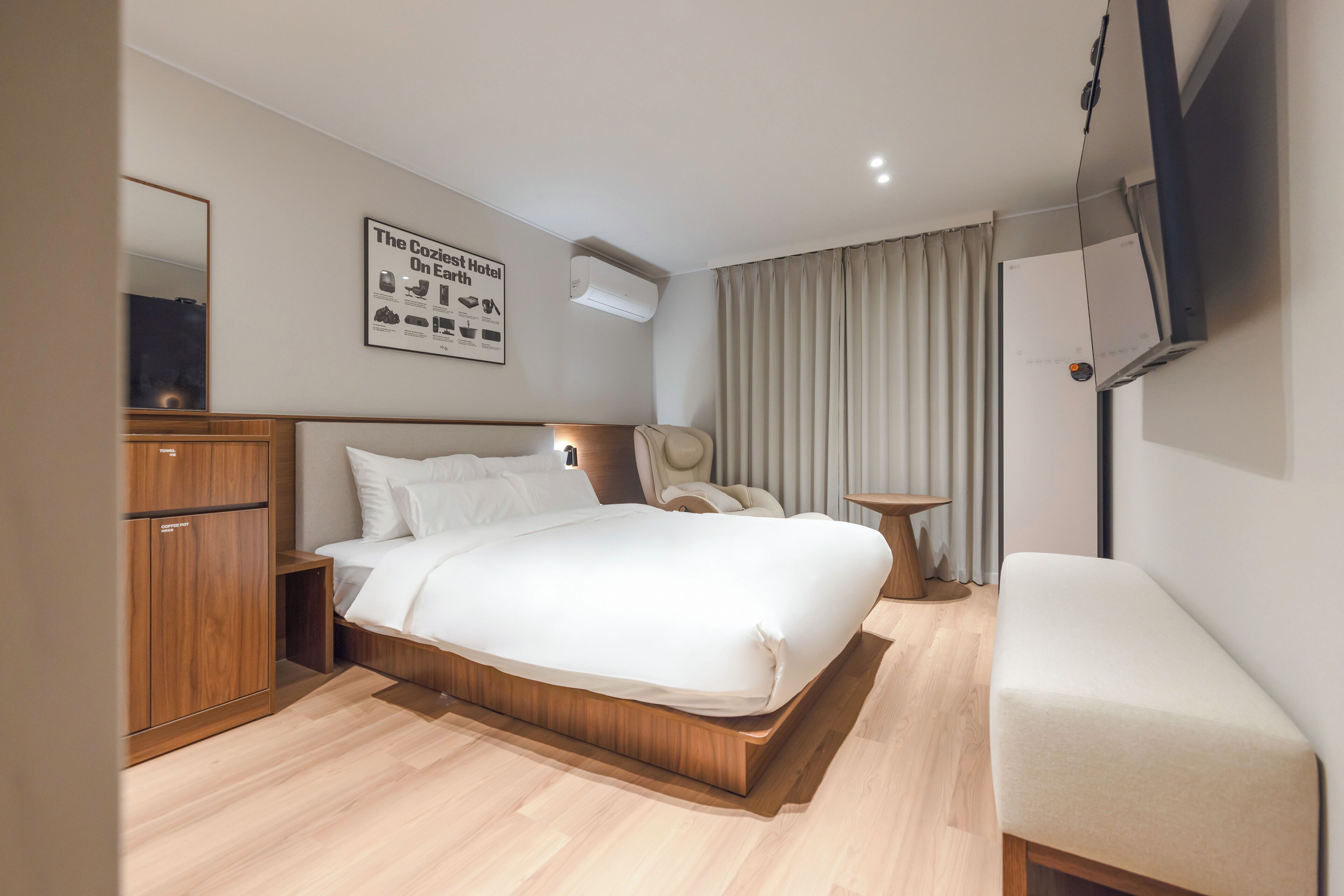A modern hotel room with a large bed, wooden wardrobe, bedside table, and a small seating area.