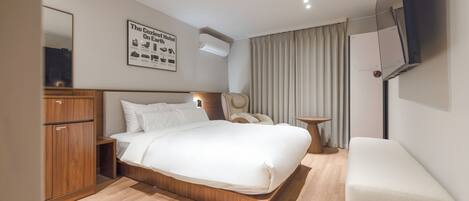 A modern hotel room with a large bed, wooden wardrobe, bedside table, and a small seating area.