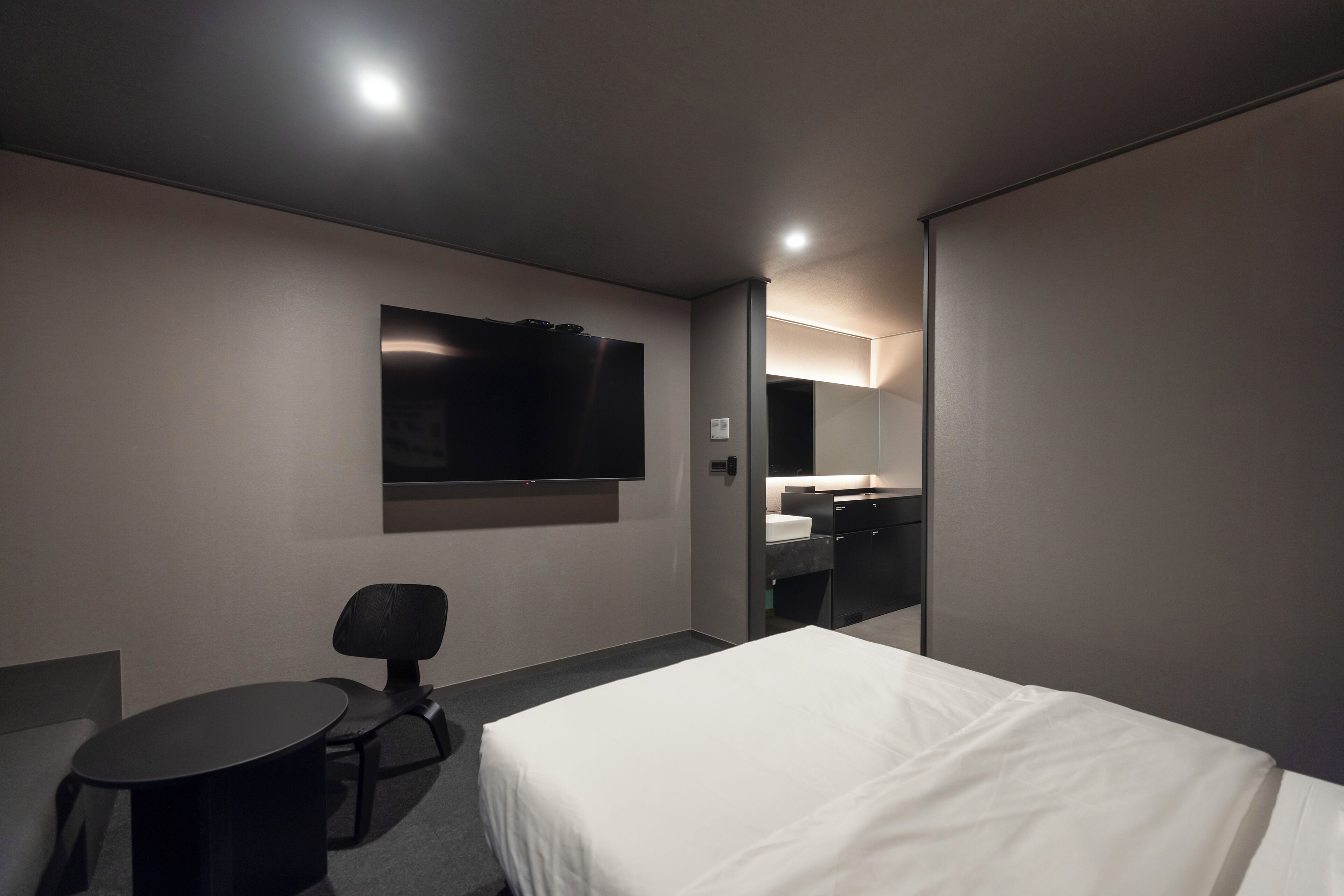 Ein modernes Hotelzimmer mit einem Bett, einem Schreibtisch, einem Sessel, einem an der Wand montierten Fernseher und einem durch eine offene Tür einsehbaren Badezimmer.