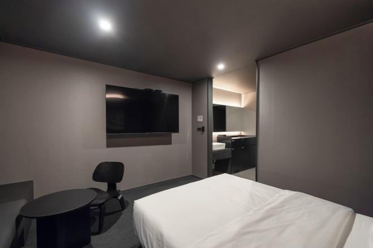 Een hotelkamer met een enkel bed, een bureau, een stoel, een aan de muur bevestigde televisie en een badkamer zichtbaar door een geopende deur.