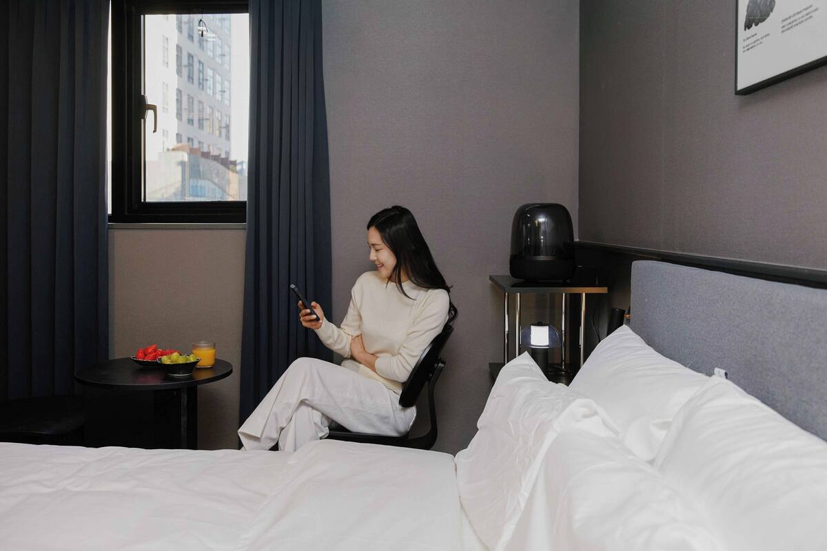 Una habitación de hotel con una cama, una mesita con un frutero y un vaso de zumo de naranja, una silla y una ventana con vistas a edificios.