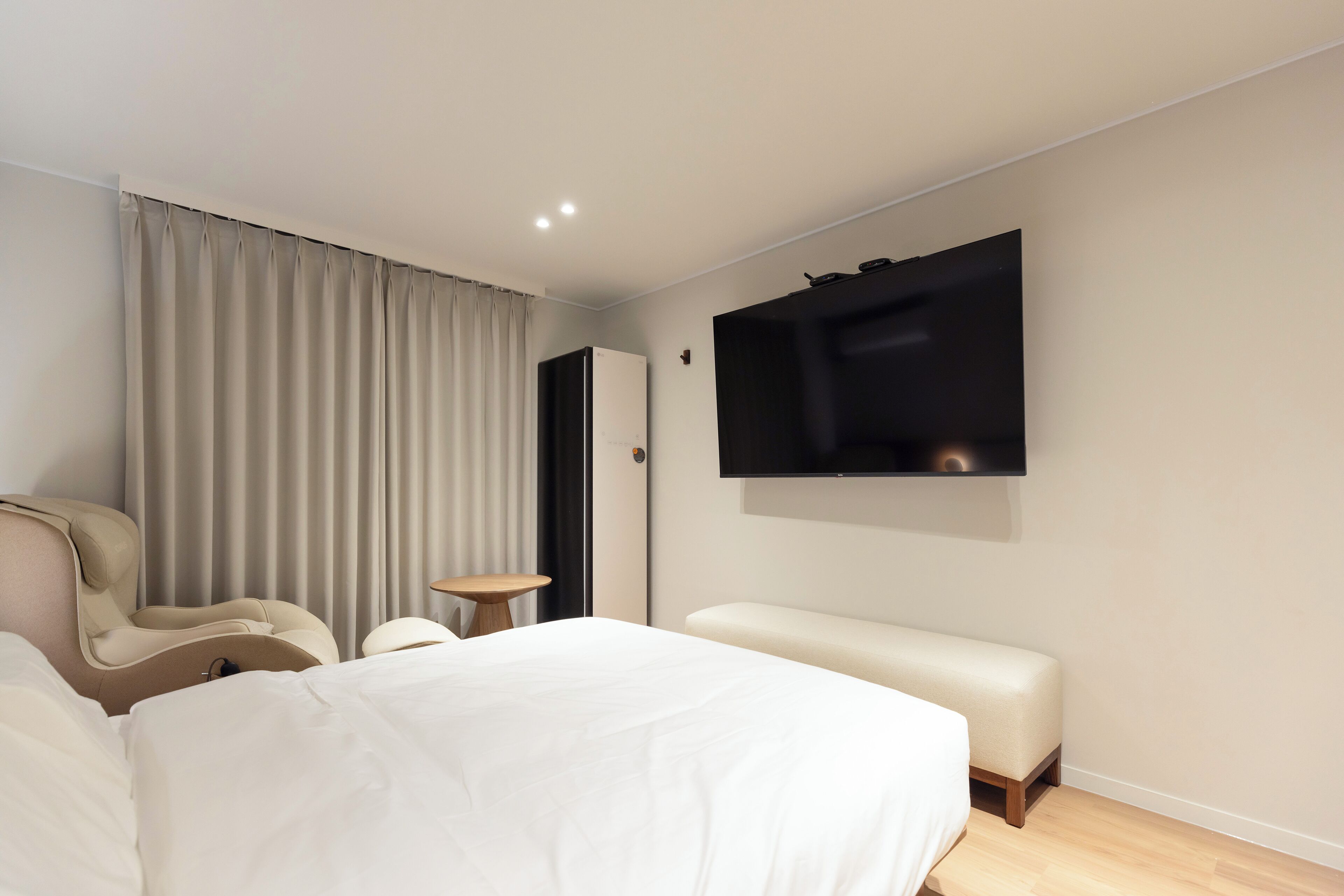 A modern hotel room with a large bed, a seating area, a flat-screen TV mounted on the wall, and a large window with curtains.
